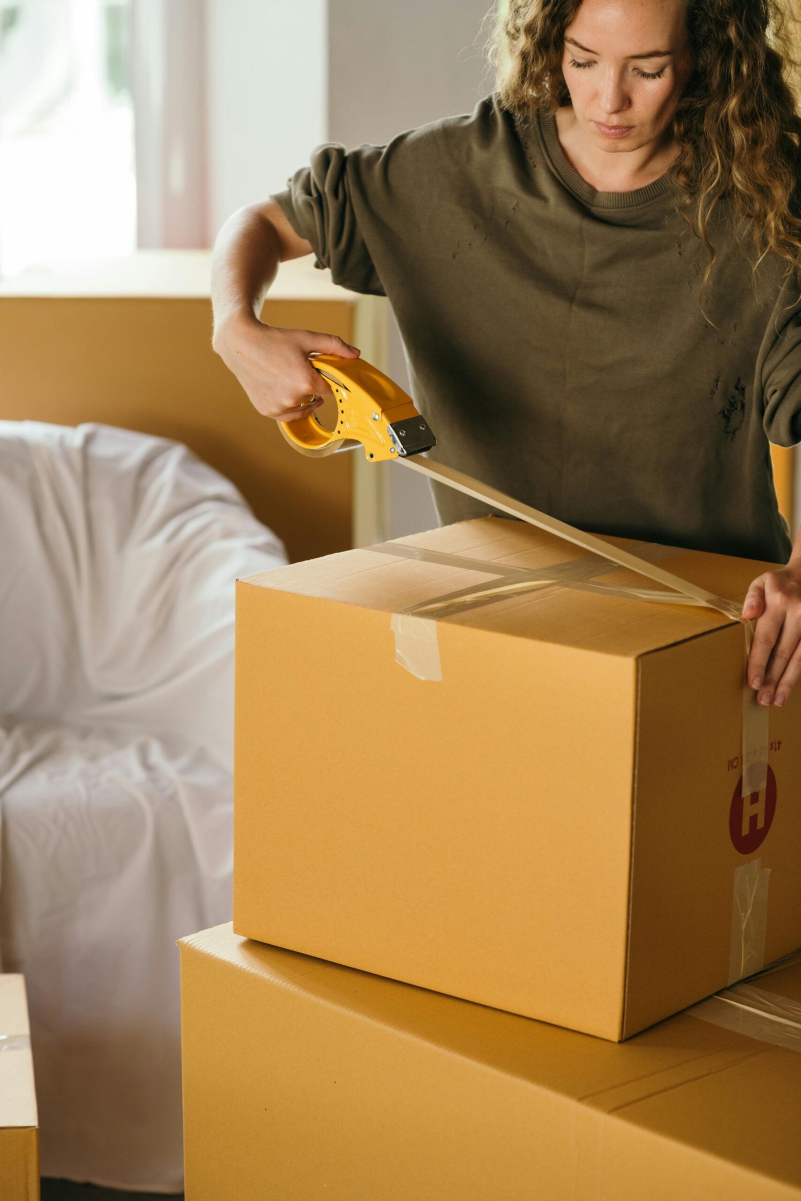 HOME 首頁 Crop female wearing casual clothes standing while packing carton boxes with scotch tape in modern apartment during preparing for relocation