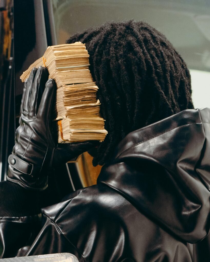 Person holding a thick stack of cash against their ear, wearing gloves and leather jacket.