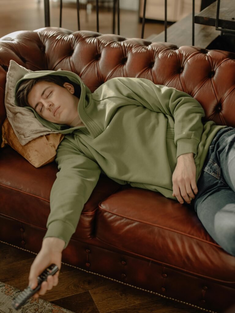A young man in a green hoodie is sleeping on a brown leather sofa, holding a remote control.