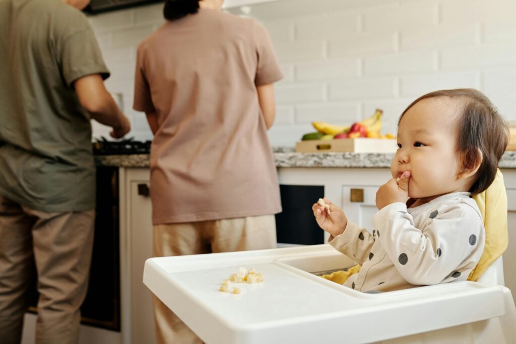 Cute toddler sits in a high chair enjoying a meal, while parents cook in the background of a modern kitchen.