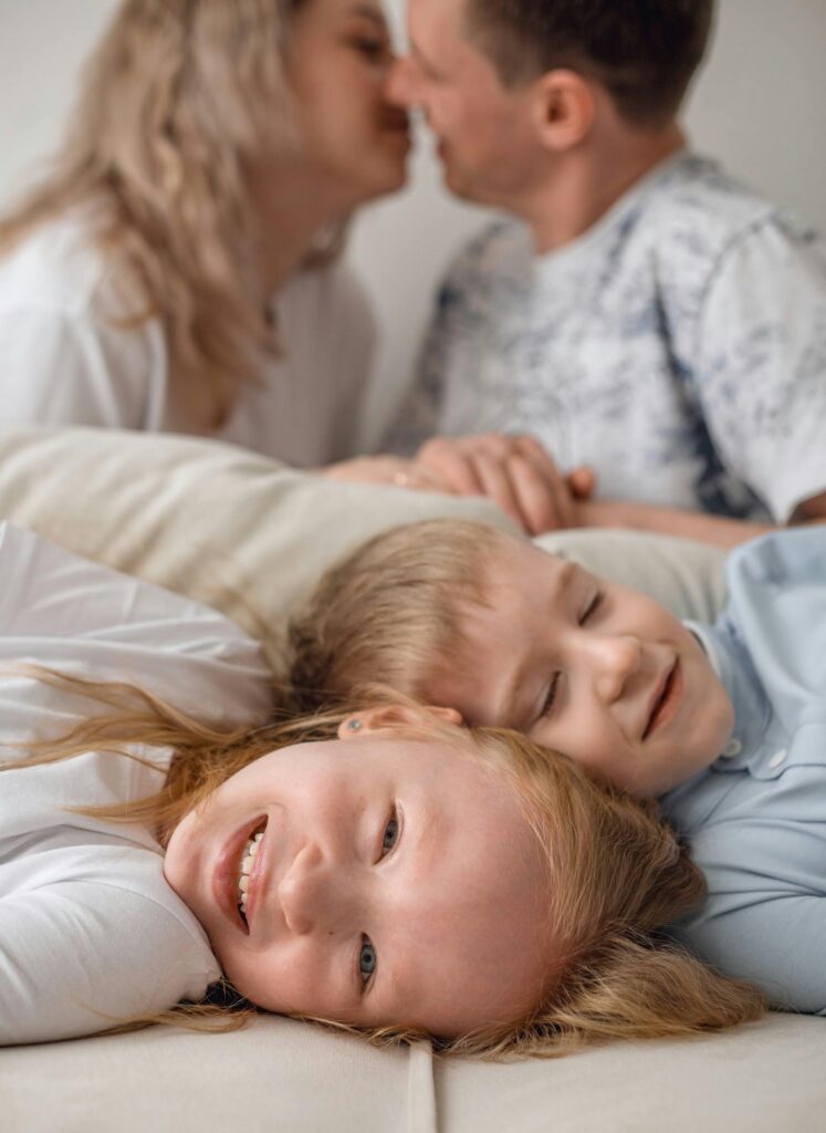 Happy children lying on couch as parents kiss in background, joyful family moment.