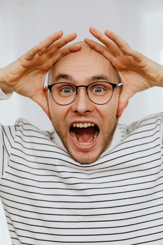 A young man with glasses screams with hands on head, showing intense emotions.