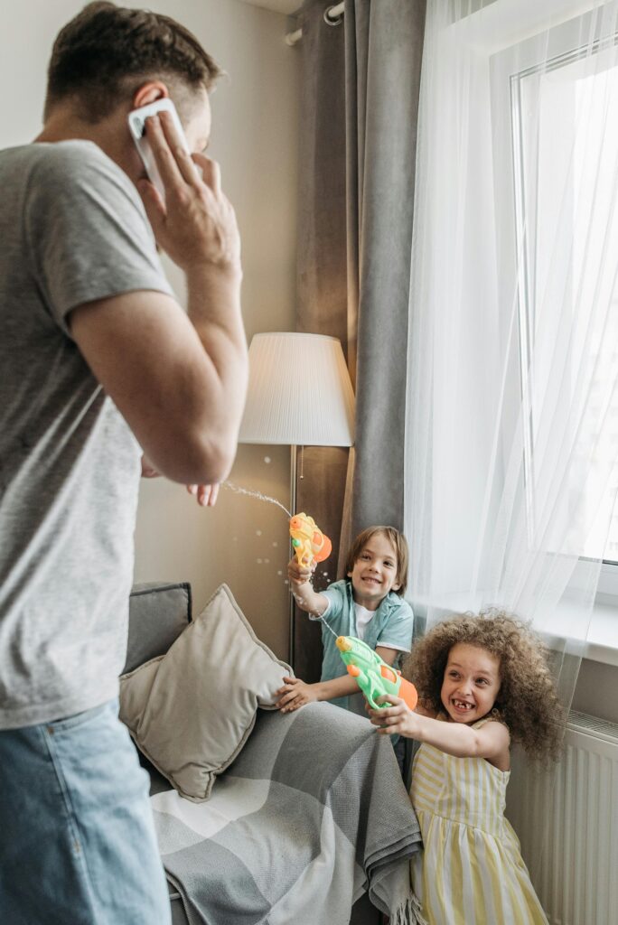 Kids playfully spraying water guns indoors while dad is on the phone.